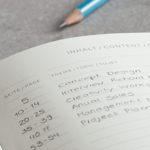 An open planner or notebook page with handwritten page numbers and German labels, shown on a gray textured surface with a blue pencil blurred in the background.
