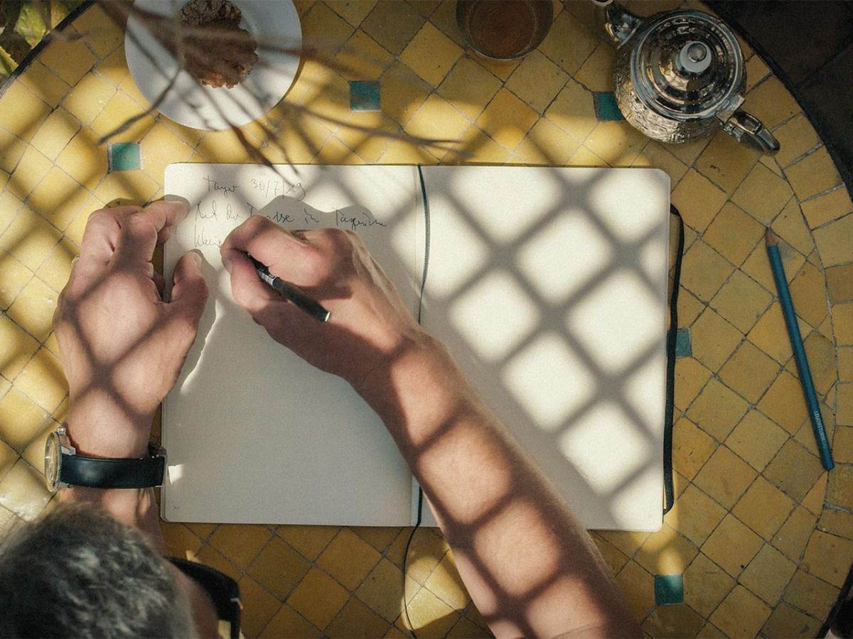 Person writing on a notepad with sunlight casting shadows on a tiled floor