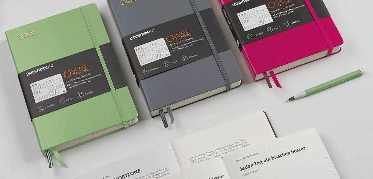 Three notebooks in green, gray, and pink with a pen on a white background