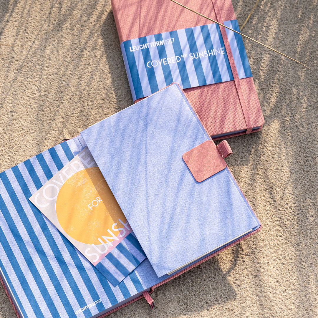 Three striped notebook covers arranged on a sandy surface in natural sunlight: one blue-and-white striped cover with a light blue flap and pink tab, one pink cover with a blue-and-white striped band and white text reading 'LUCKILY 2027 COVERED SUNSHINE,' and one blue-and-white striped cover with a yellow circular insert.