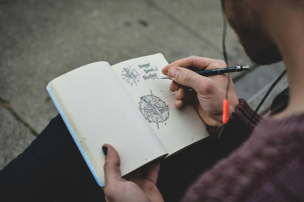 Person drawing in a small notebook with a pen on a sidewalk.