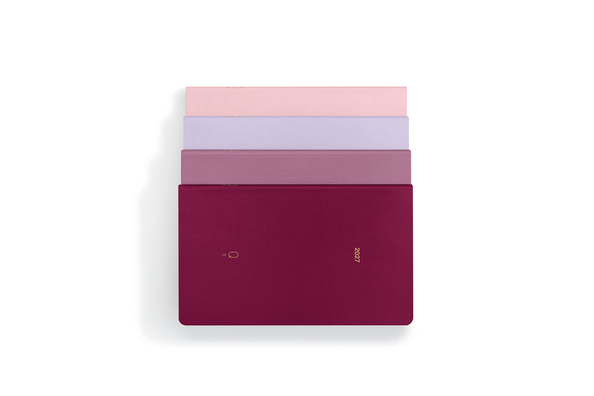 Four slim rectangular notebooks or journals stacked vertically on a white background, with the top three fanned out slightly. The bottom notebook is a deep burgundy/magenta color with a small gold logo, while the others show pink, lavender, and mauve tones.