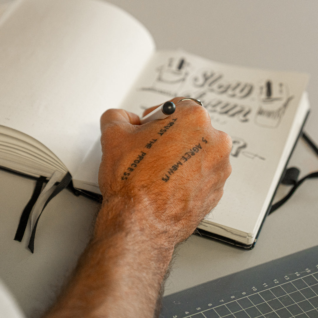 Hand with tattoos holding a pen over an open notebook on a desk.