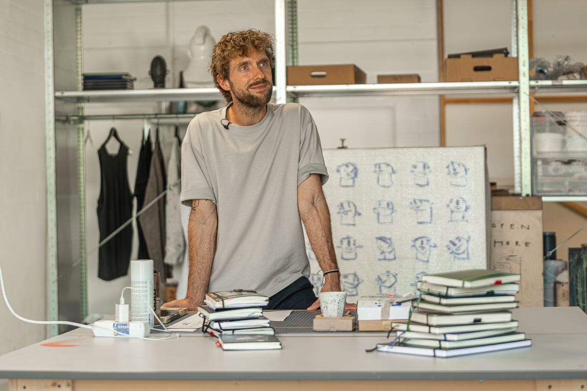 Man standing behind a table with books and artwork in a room with shelves and clothing.