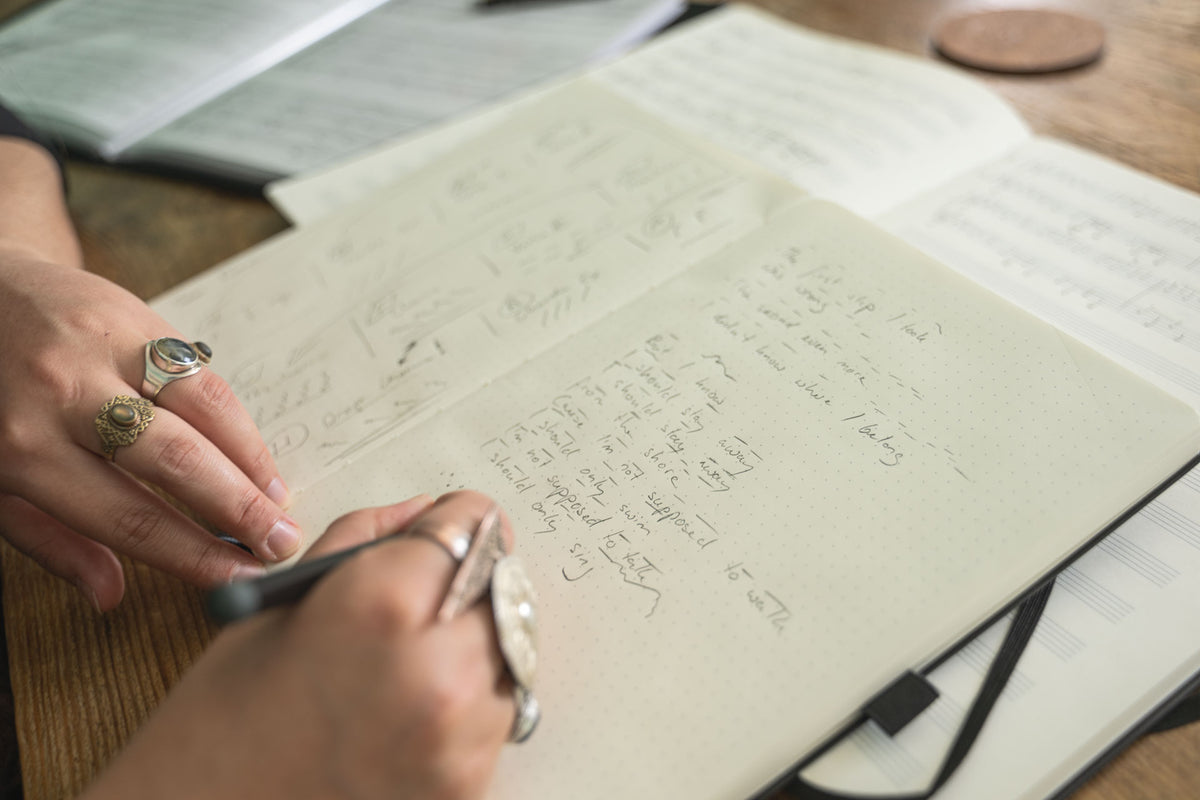 Person writing in a notebook with a pen on a wooden desk.