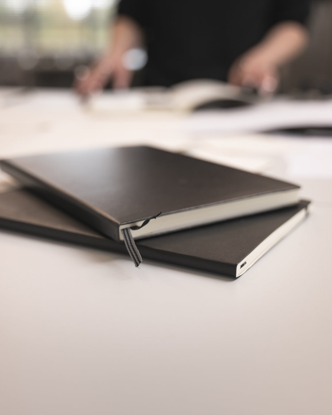 Two black notebooks on a light surface with a blurred background of people and books.