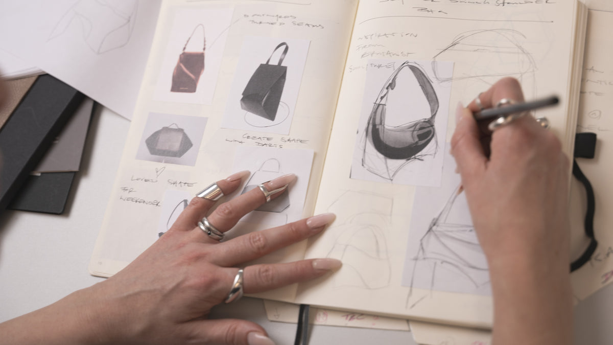Person sketching designs of handbags in a notebook with a pen.