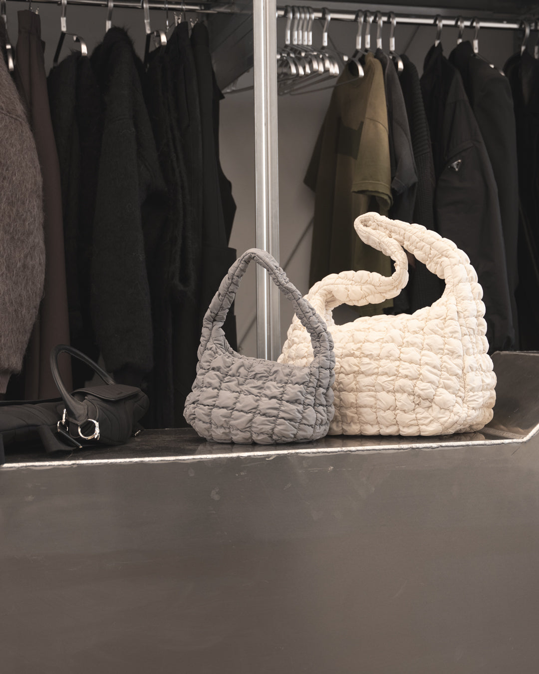 Two textured handbags, one gray and one white, on a dark surface with a clothing rack in the background.