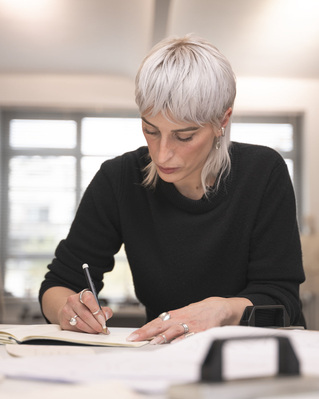 Woman with short blonde hair wearing a black sweater, sitting at a desk and writing in a notebook.
