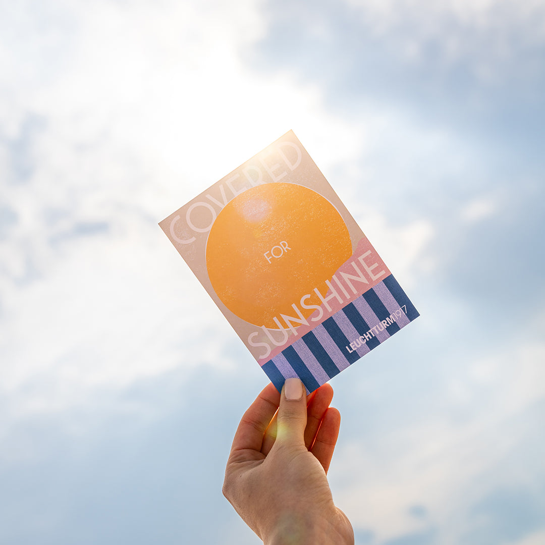 A hand holds a square card or book cover against a cloudy sky. The cover features a large yellow-orange sun graphic with the words 'COVERED FOR SUNSHINE' and 'LIGHTYEAR 7' in blue and white text.