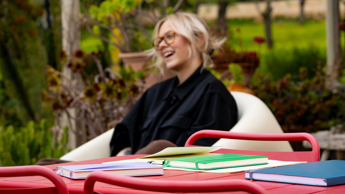 Person sitting outdoors with books on a table, surrounded by nature