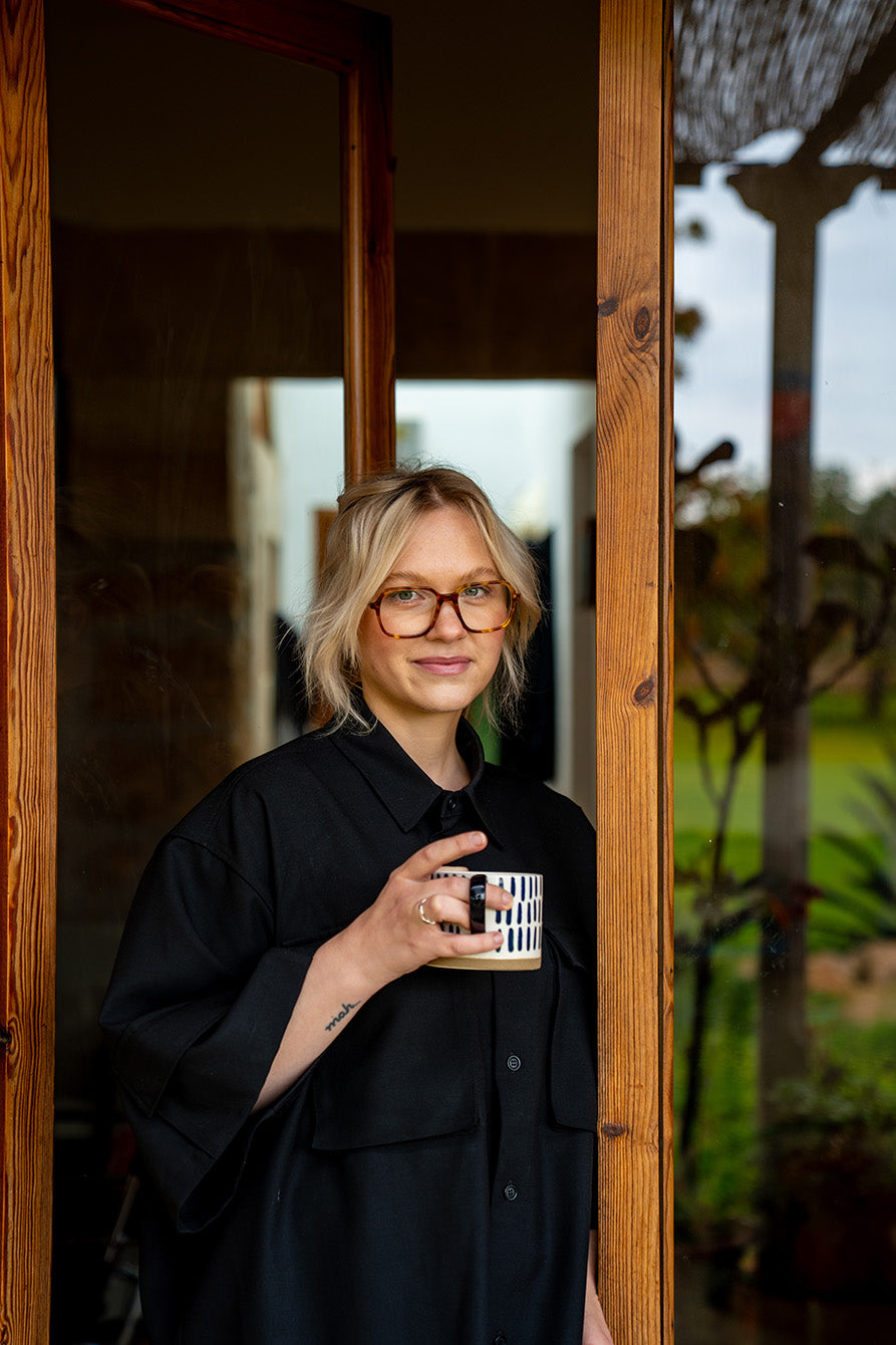 Woman holding a mug with a scenic design, standing in a wooden doorway.