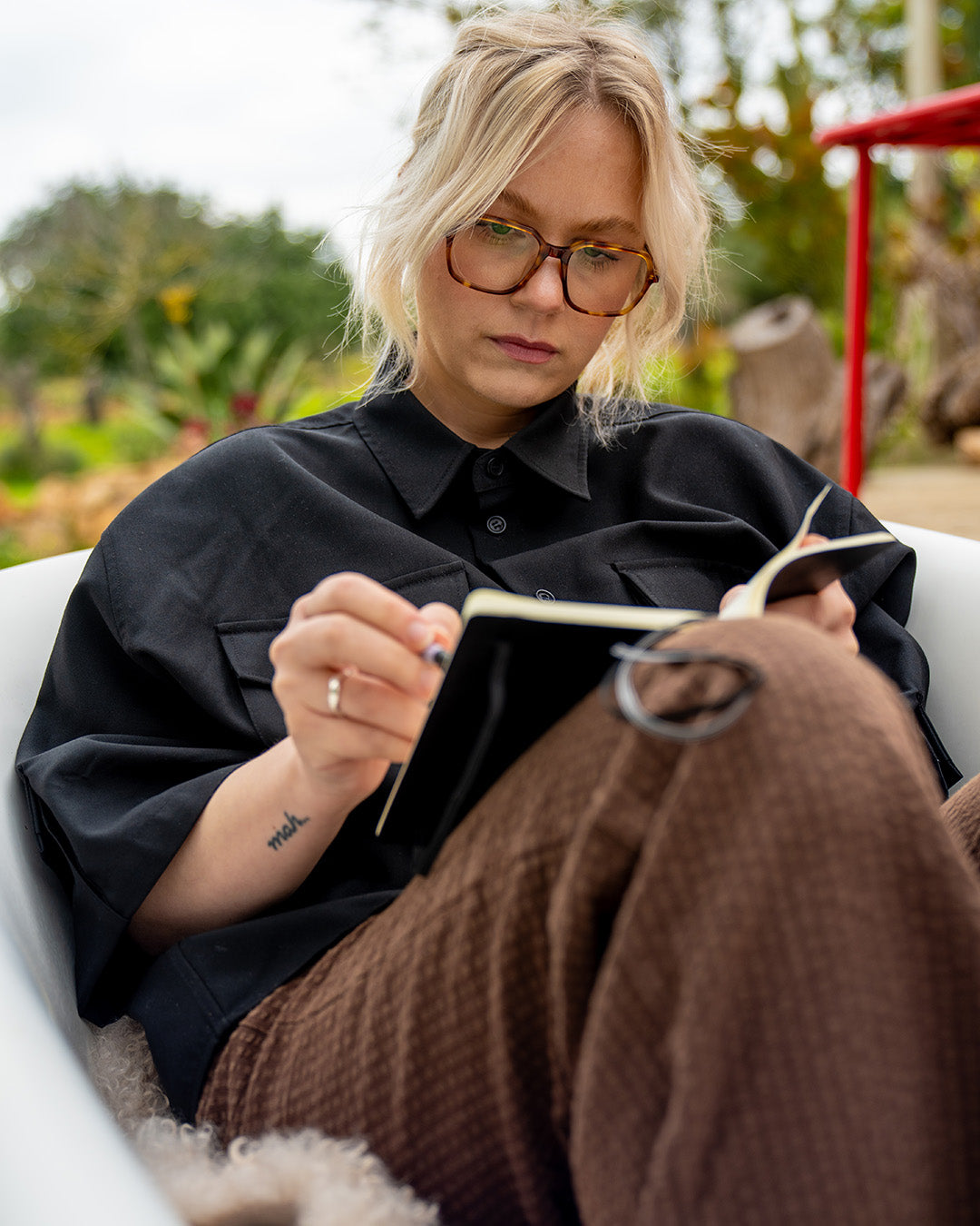 Person sitting outdoors, holding a book, wearing glasses and a black shirt.