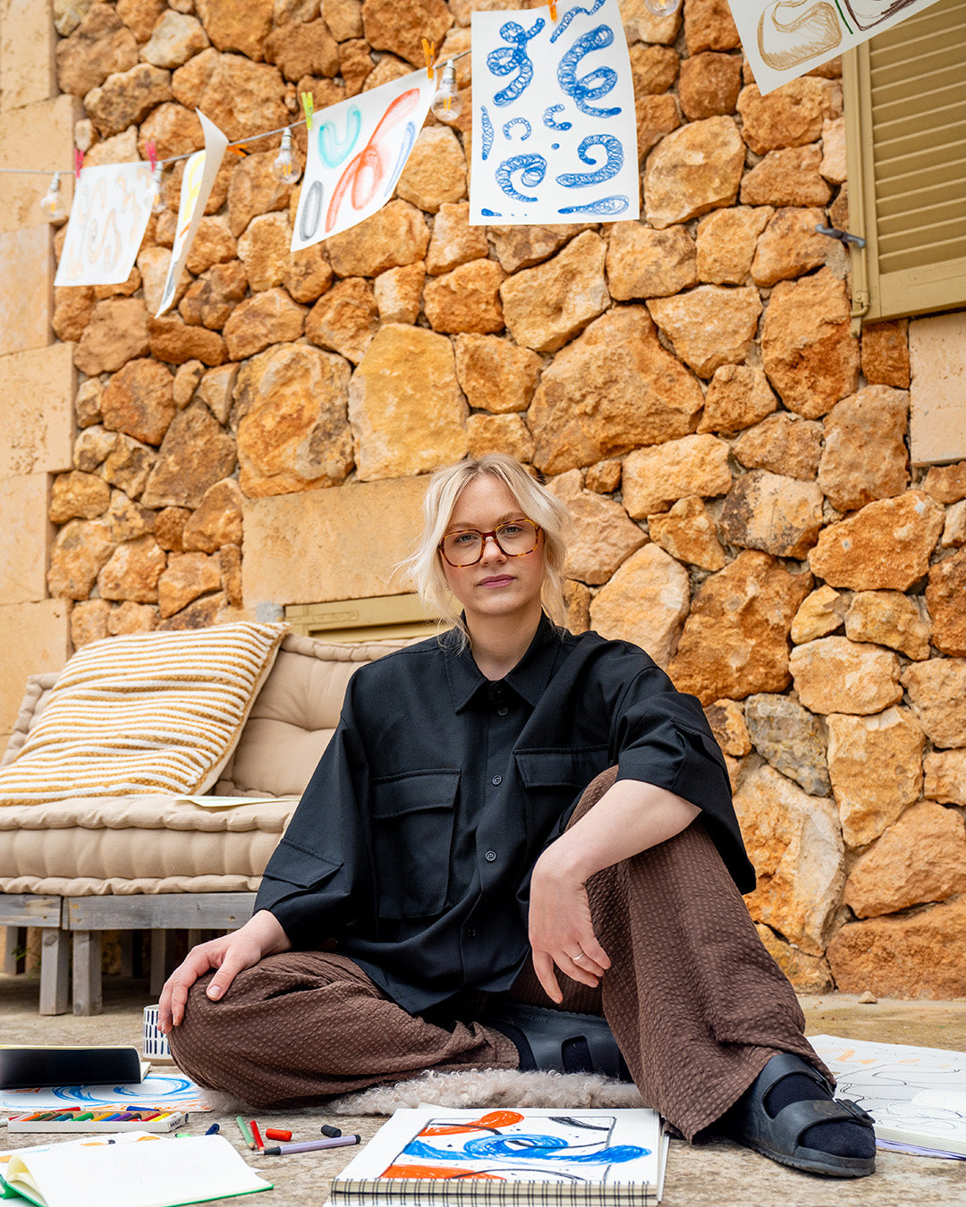 Person sitting on a stone wall with art supplies, wearing glasses and a black shirt.