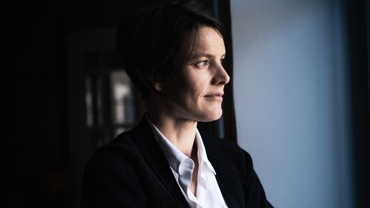 Woman in a dark jacket and white shirt looking out a window with a blurred background