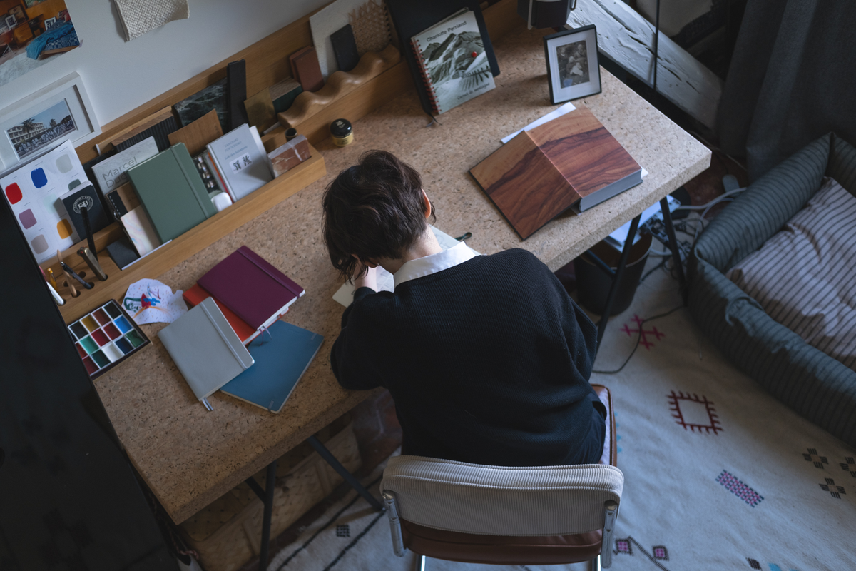 Person sitting at a desk with books and art supplies in a home office setting.