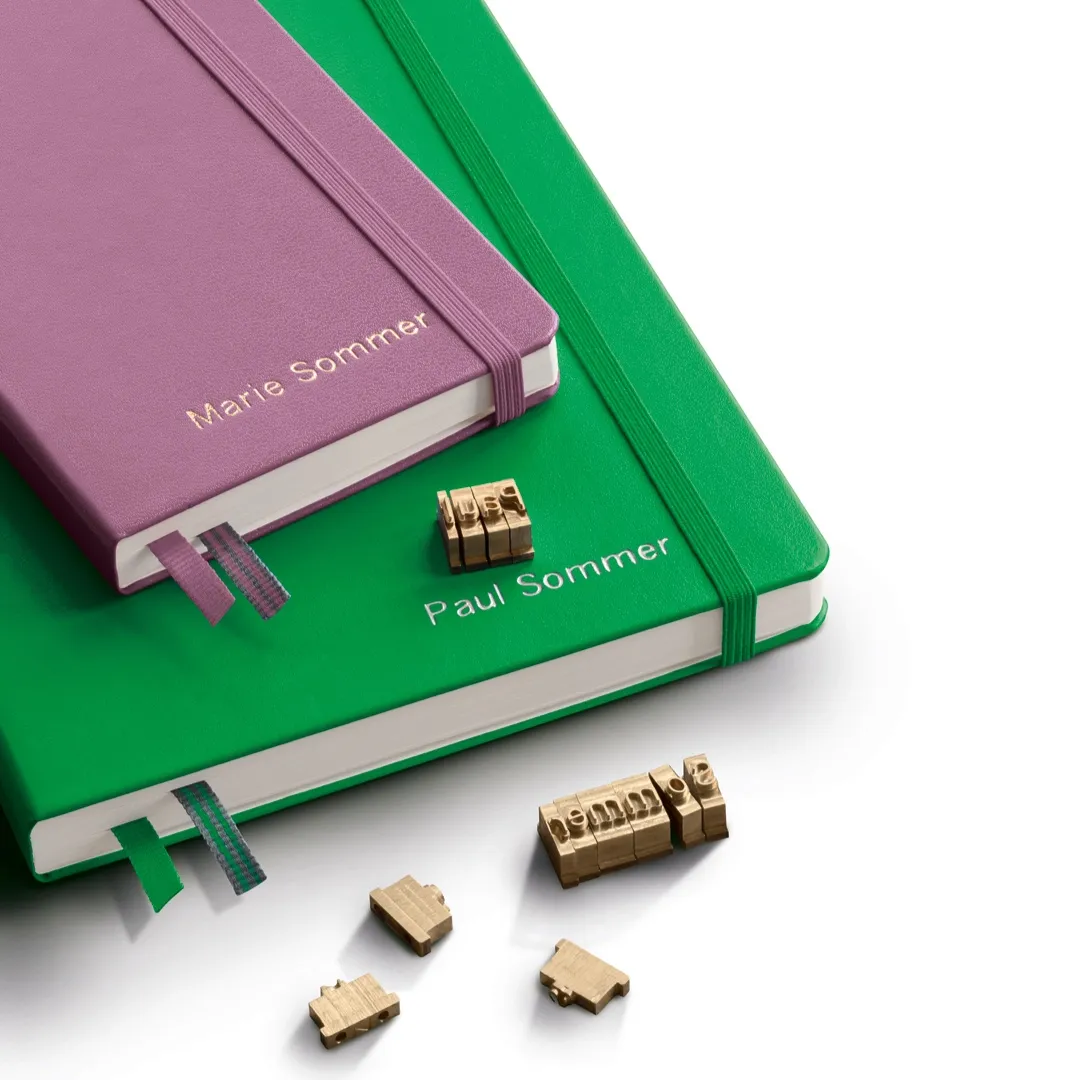 Green notebook with a purple cover featuring engraved names, accompanied by small gold lettering on a white background.