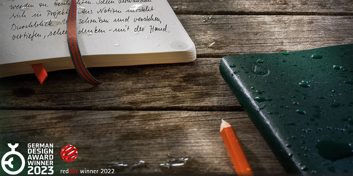 Open book with handwritten text on a wooden surface, next to a green notebook and orange pencil.