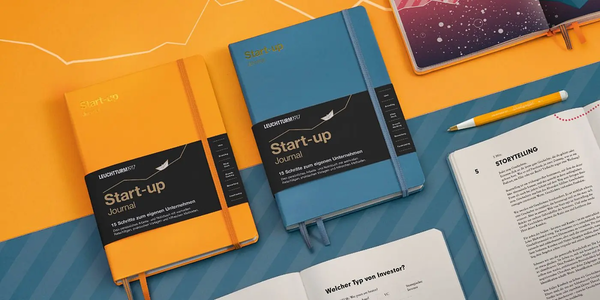 Two 'Start-up Journal' notebooks on a colorful background with a pen and paper.