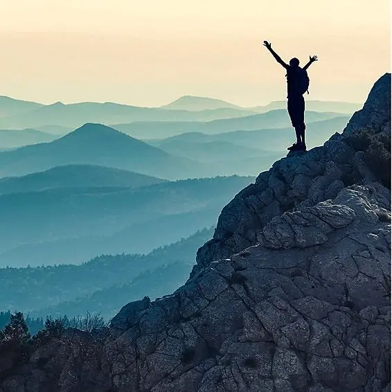 Person standing on a mountain peak with arms raised, overlooking a scenic view of mountains.