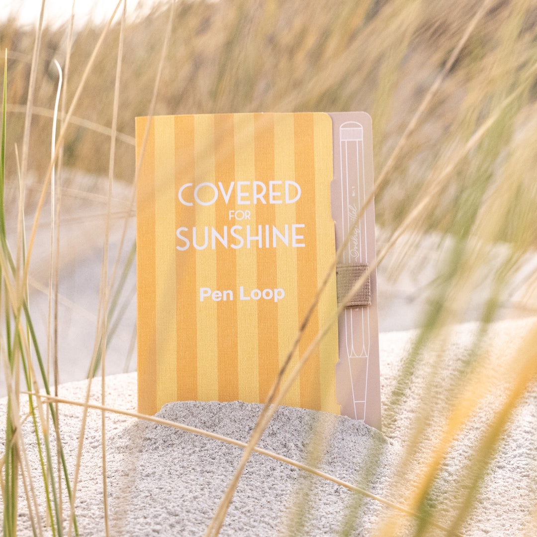 A pen loop notebook with yellow and orange vertical stripes, standing upright in sand with beach grass in the background. The cover reads 'COVERED FOR SUNSHINE' and 'Pen Loop'.
