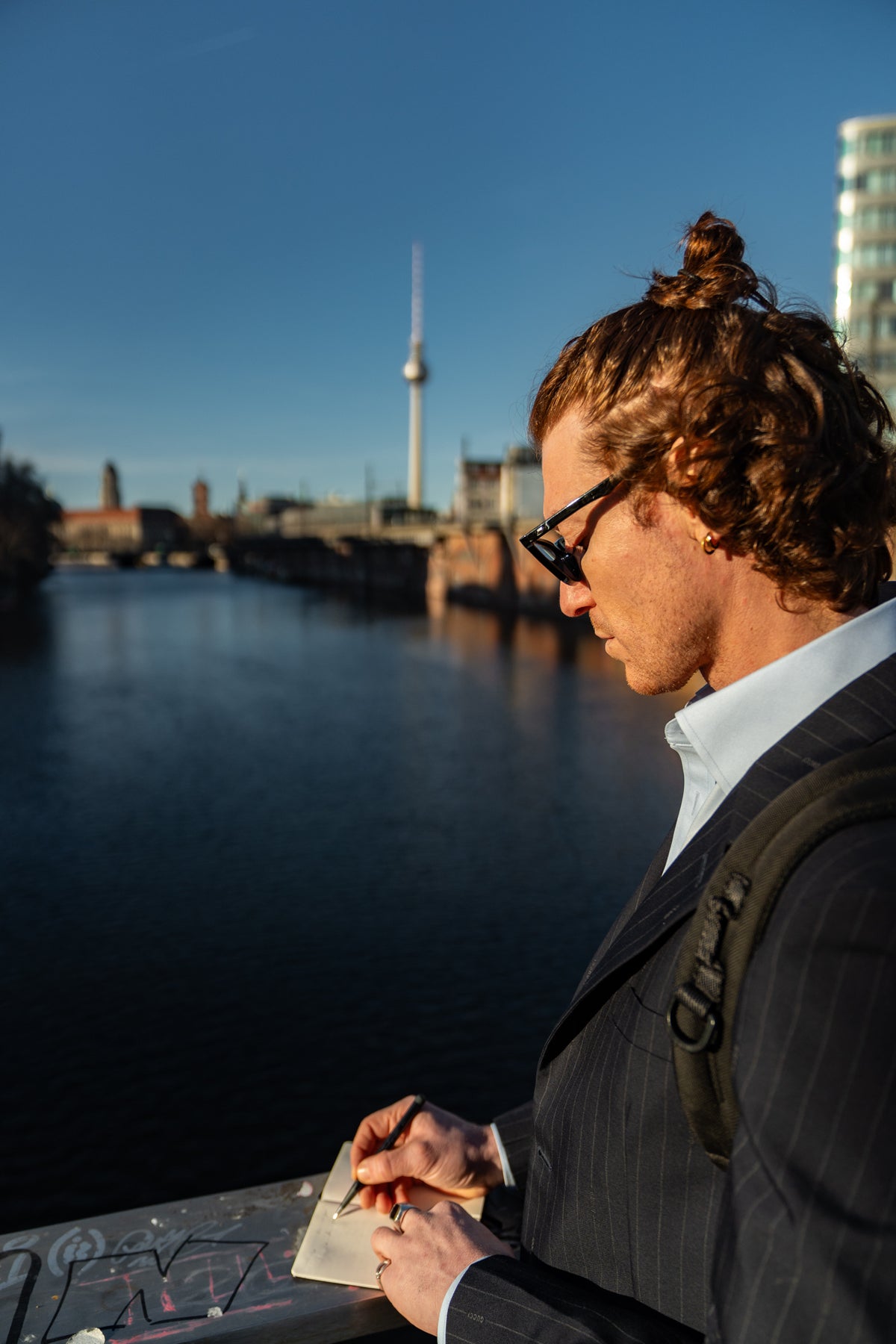 Person with braided hair and a backpack standing by a waterfront with a city skyline in the background.