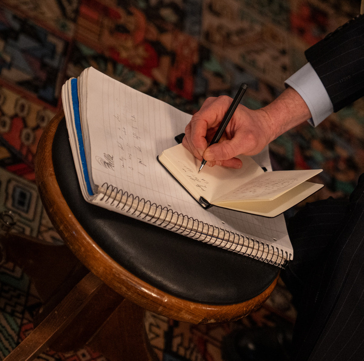 Person writing in a notebook on a bar stool with a patterned rug in the background