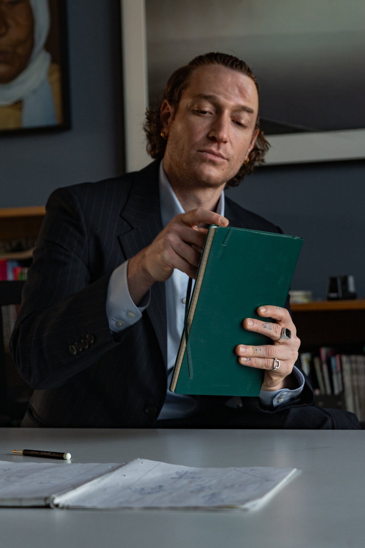 Man in a suit reading a green book at a desk with papers and a pen.