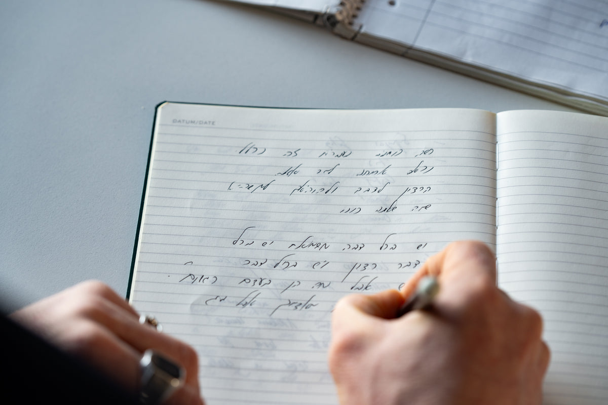 Person writing in a notebook with a pen on a light surface.