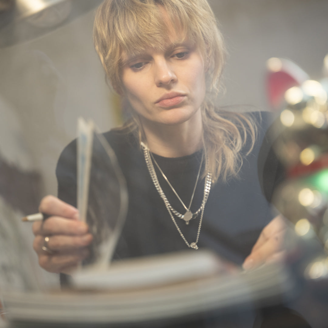 Person with long blonde hair holding a cigarette in a dimly lit room.