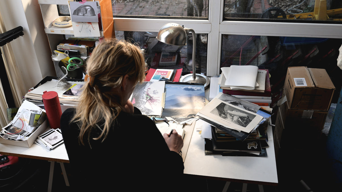 Person sitting at a cluttered desk with various items including books and a camera.