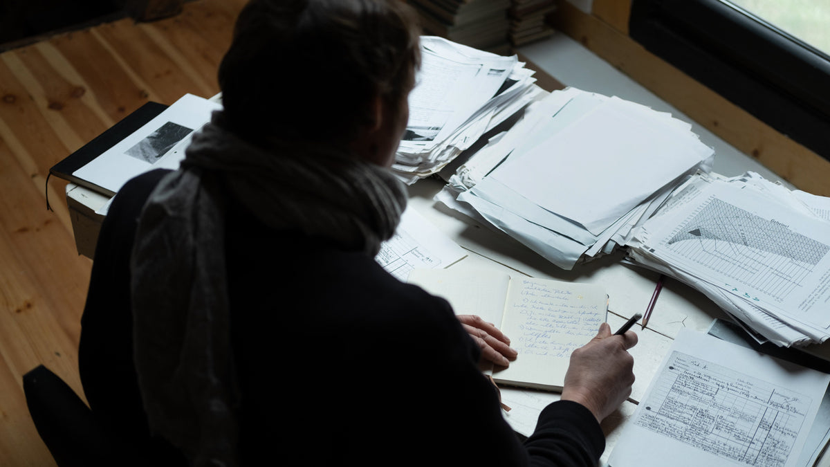 Person sitting at a desk with stacks of papers, using a pen.