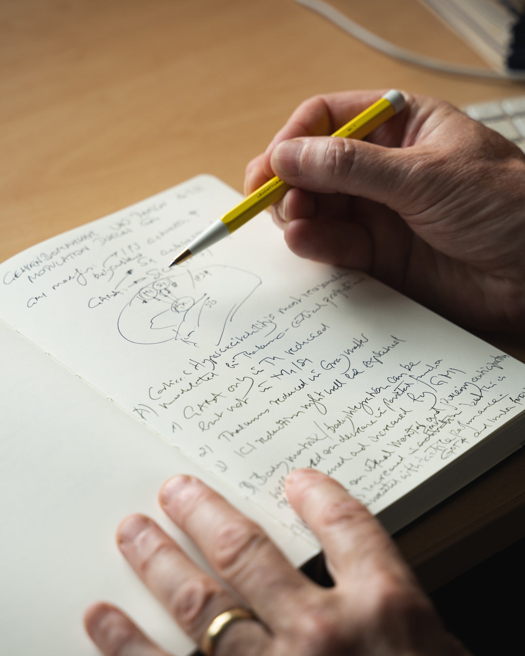 Hand writing in a notebook with a yellow pen on a wooden surface.