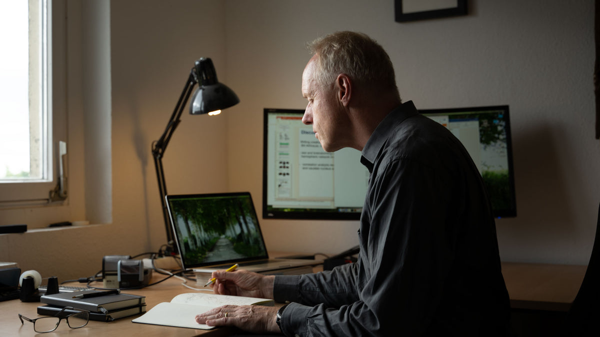 Man sitting at a desk with multiple computer monitors and a lamp in a home office setting.