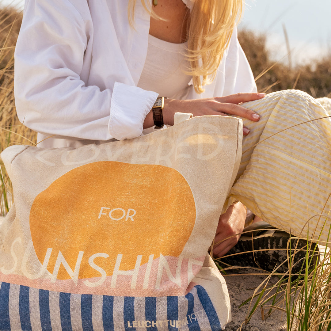 A beige canvas tote bag with a large yellow circle and the words 'FOR SUNSHINE' in white, carried by a woman in a white shirt in a sunny outdoor grassy setting.