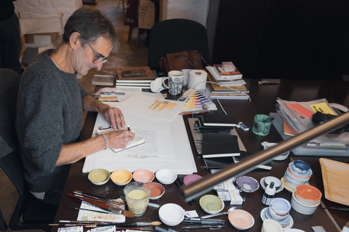 Artist sitting at desk surrounded by paint supplies writing in a notebook
