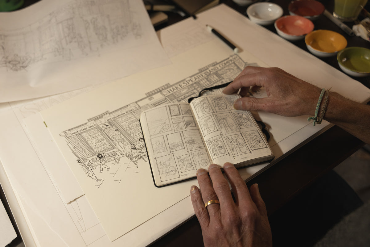 Person working on a detailed design in a notebook with paints in the background