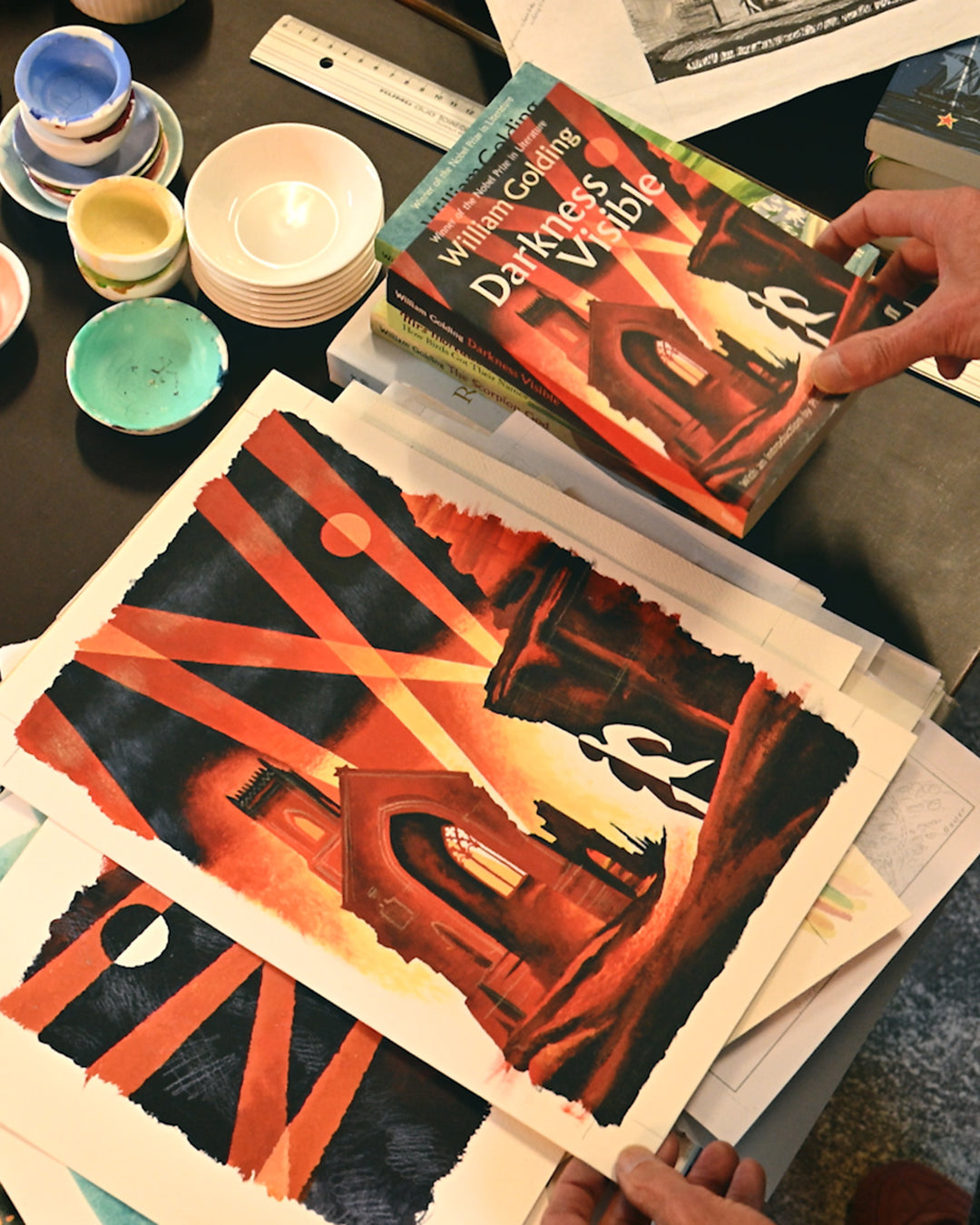 Person holding a book titled 'Darkness Visible' next to art prints on a table.