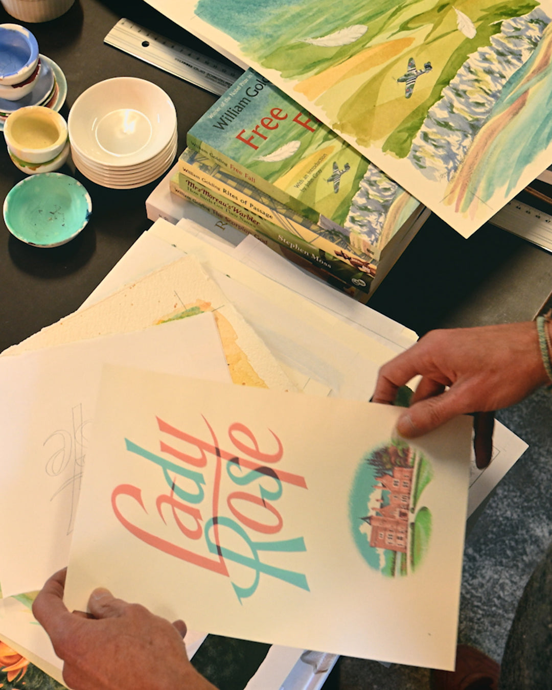 Person holding a sheet of paper with 'Lady Rose' printed on it, surrounded by art supplies.
