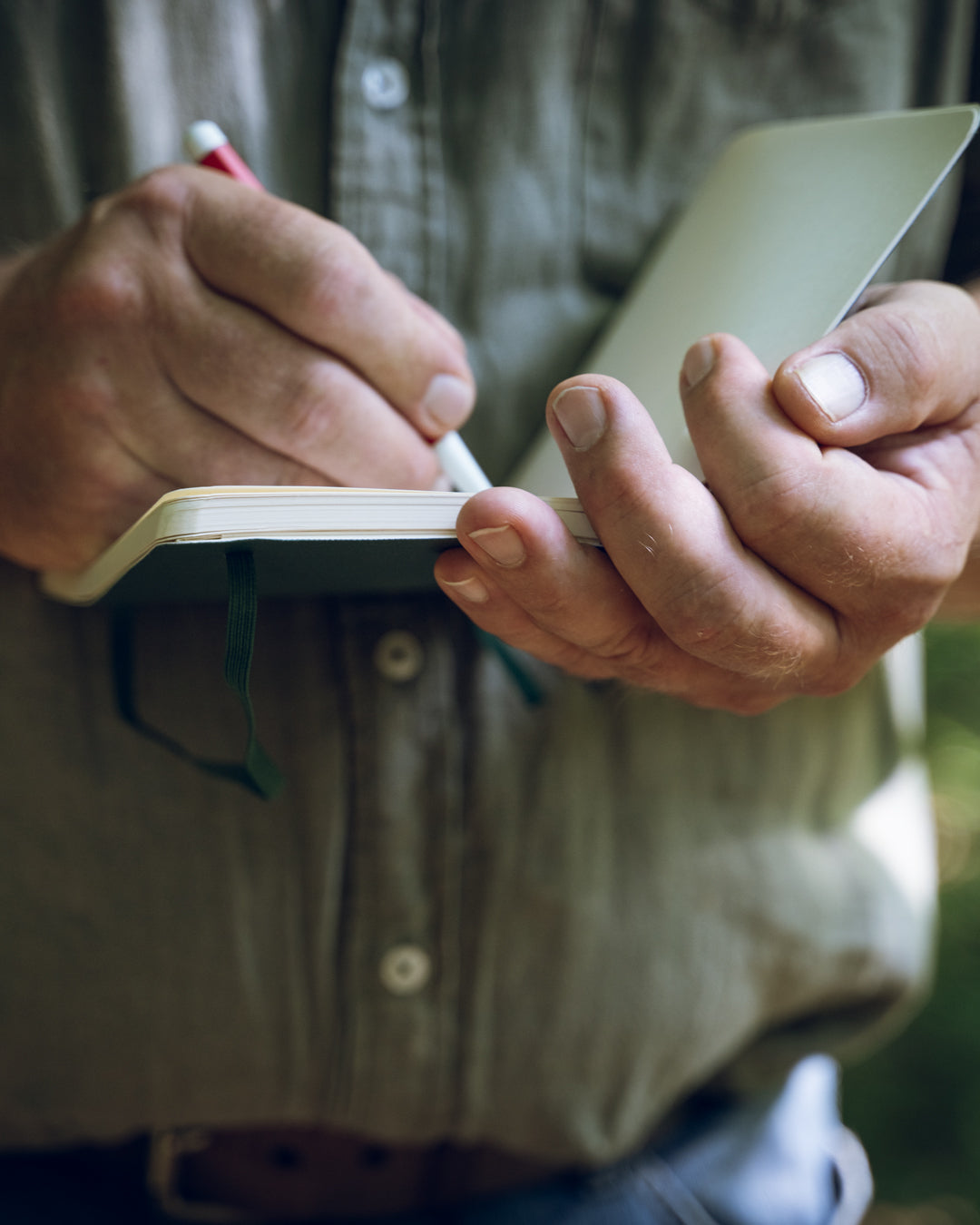 Person writing in a small notebook with a pen, wearing a brown shirt.