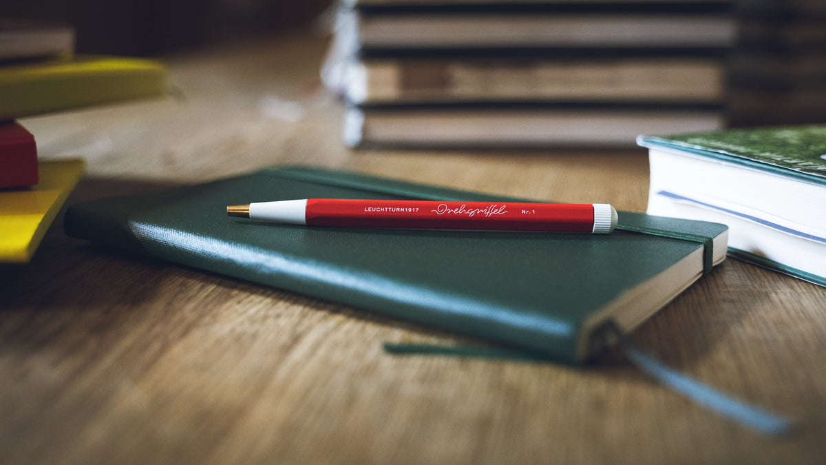 Green notebook with a red pen on a wooden surface, with books blurred in the background
