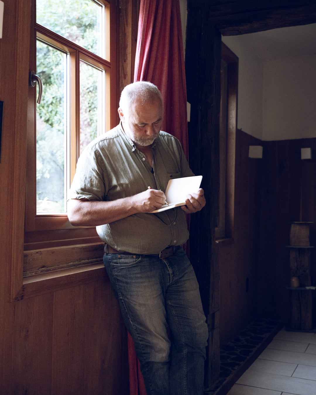 Man writing on a piece of paper by a window in a room with wooden walls.