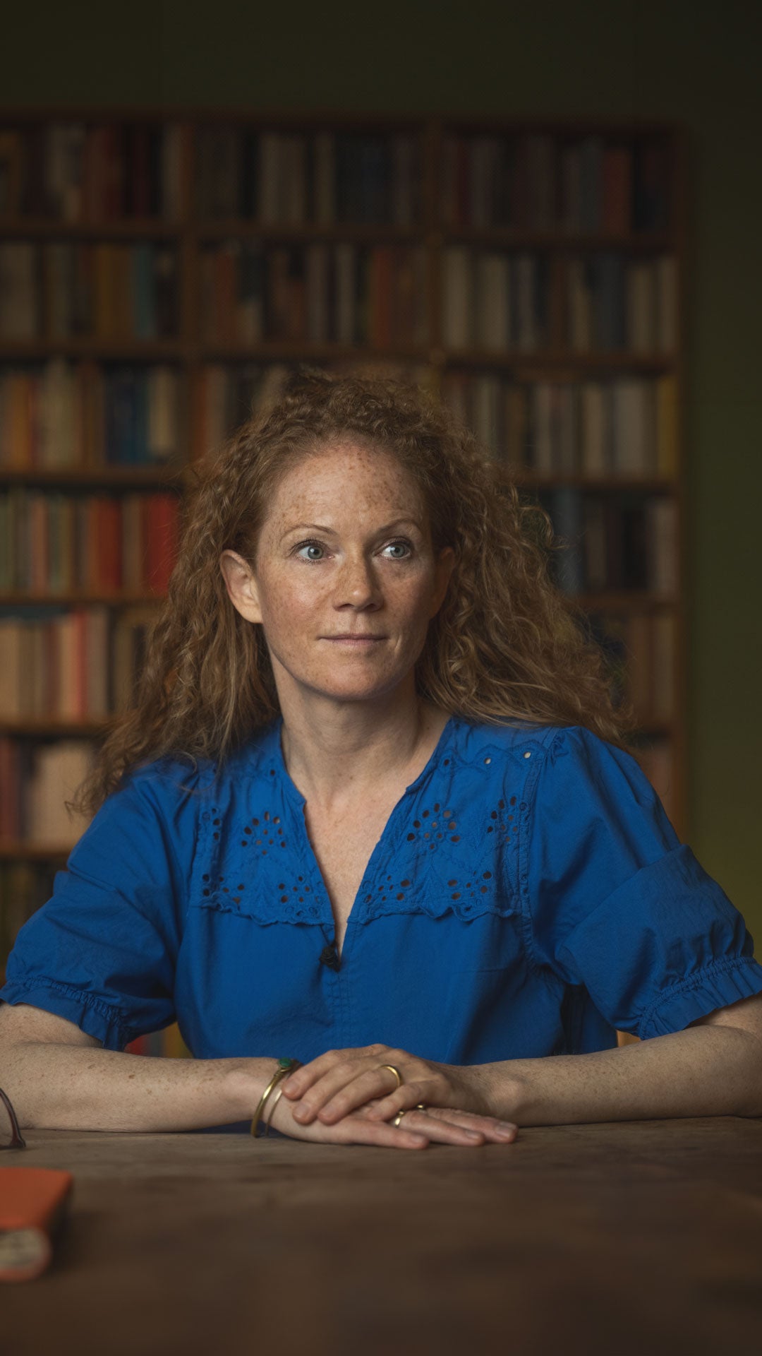 Woman in a blue shirt sitting at a table with a bookshelf in the background