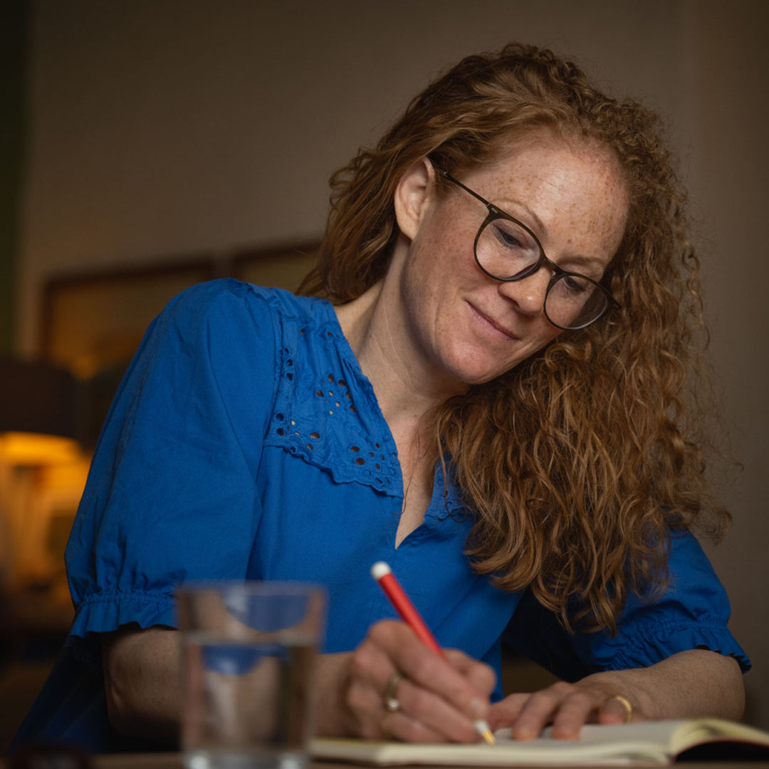 Woman in a blue shirt writing in a notebook with a red pen, surrounded by glasses on a table.