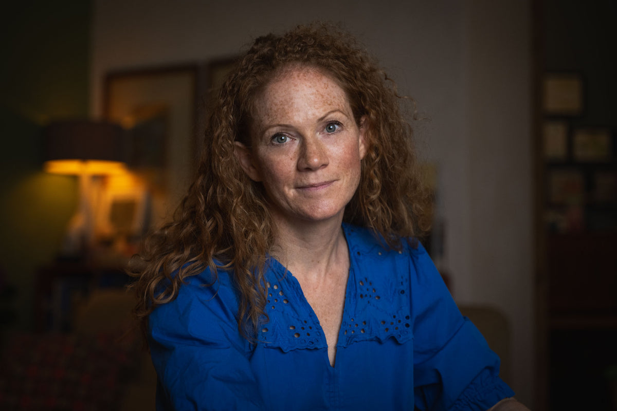 Woman with red hair wearing a blue shirt in a dimly lit room.
