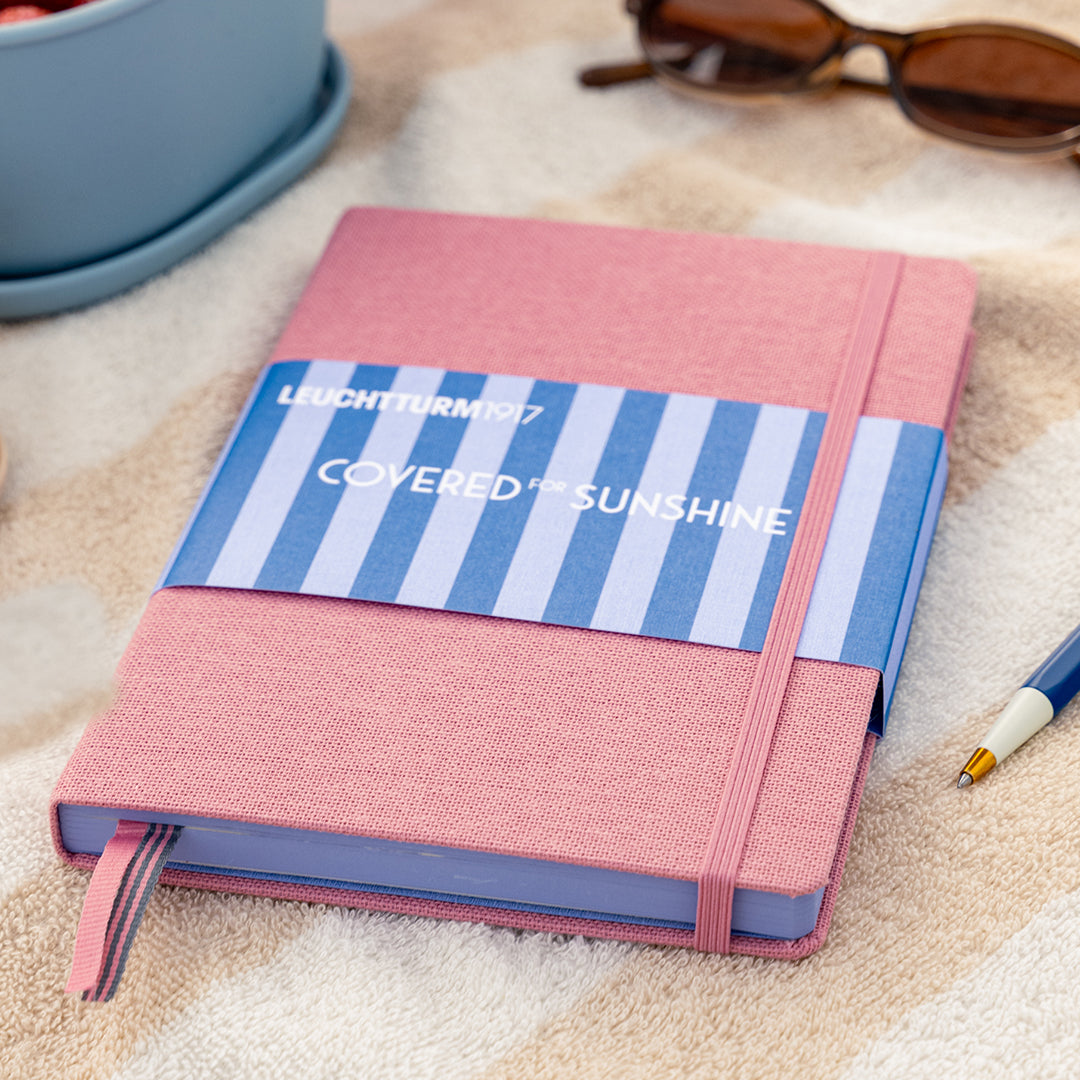 Pink notebook with blue striped label on a textured surface with sunglasses and a pen.